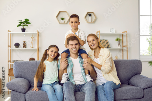 Together at home. ortrait of loving young couple and their children posing together on sofa in living room. Happy Caucasian amily of four is smiling joyfully looking at camera. photo