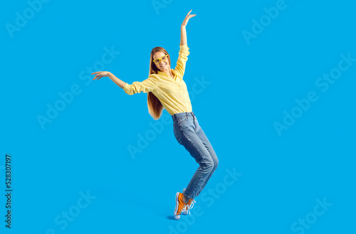 Overjoyed millennial Caucasian girl in casual wear and glasses isolated on blue studio background dancing. Happy funny young woman have fun make dancer moves. Hobby and entertainment.
