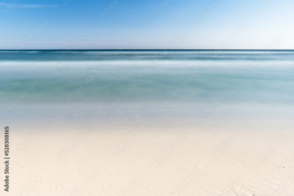 Long exposure of a beach on the Gulf of Mexico. The water is blue with golden sand and a blue sky. 