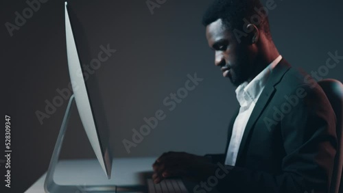 African-American male dark-skinned businessman working at night at a computer monitor in his office against a gray background against a wall