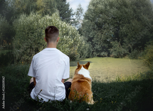 A teenage boy and a corgi dog sit with their backs to the pond next to each other watching dreaming, love for animals