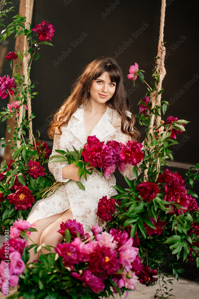 a beautiful young woman in a white dress sits on a swing in peony flowers. a beautiful young woman in a white dress sits on a swing in peony flowers.
