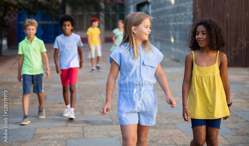 Happy blonde preteen girl walking with her african american girl playmate along city street on summer day ..