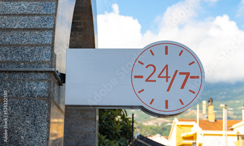 A white banner with the image of a clock signifying the round-the-clock operation of the institution against the background of a blue sky. twenty-four by seven round-the-clock work signage