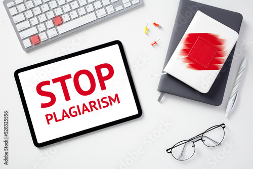 Stop plagiarism concept. Flat lay digital tablet with text STOP PLAGIARISM on screen, keyboard with lighted keys control c, paper notebooks, glasses on white desk.