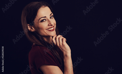 Beautiful young business toothy smiling woman thinking and looking happy in burgundy blouse and with red lipstick on dark shadow black background with empty copy space. Portrait