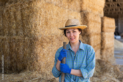 Portrait of positive young asian female farmer standing leaning on pitchfork near straw stack in hayloft, posing during working at farm