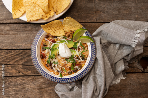 Chicken tortilla soup with sour cream, avocado, lime, tortilla chips and napkin on wooden table, from above