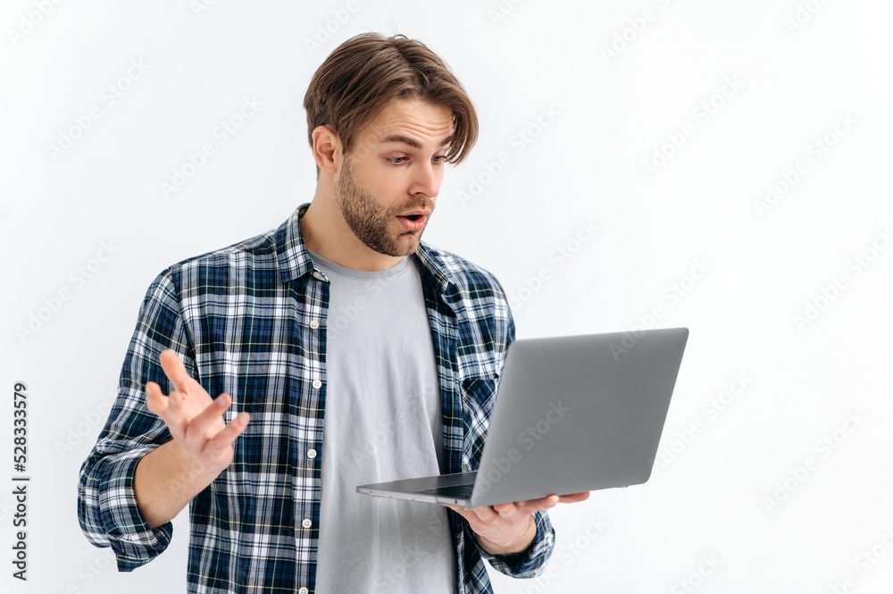 Shocked confused puzzled caucasian stylish young man, standing on a white isolated background, holds an open laptop, surprised looks at the screen, see unexpected news, message, gesturing with hand Shocked confused puzzled caucasian stylish young man, standing on a white isolated background, holds an open laptop, surprised looks at the screen, see unexpected news, message, gesturing with hand