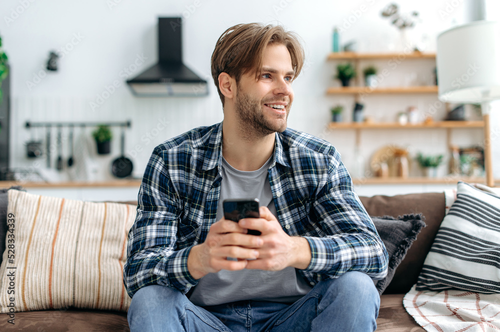 Positive handsome stylish caucasian young man, sitting with smartphone in hands on couch at home in living room, messaging online with friends, browsing websites, looks away, dreaming, smiling