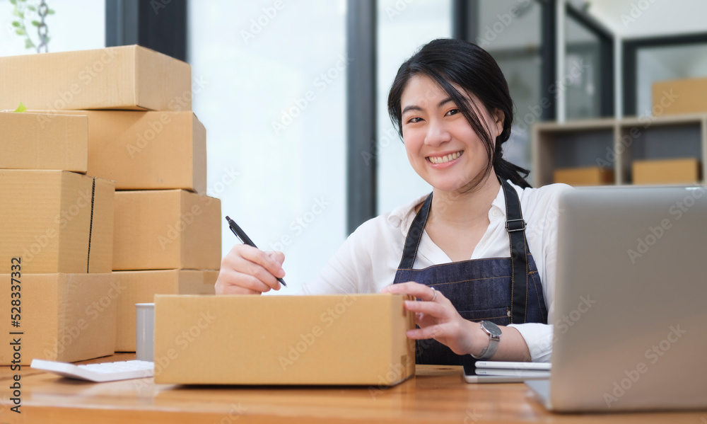 Smiling young female start up small business owner writing address on cardboard box. Smiling young female start up small business owner writing address on cardboard box.