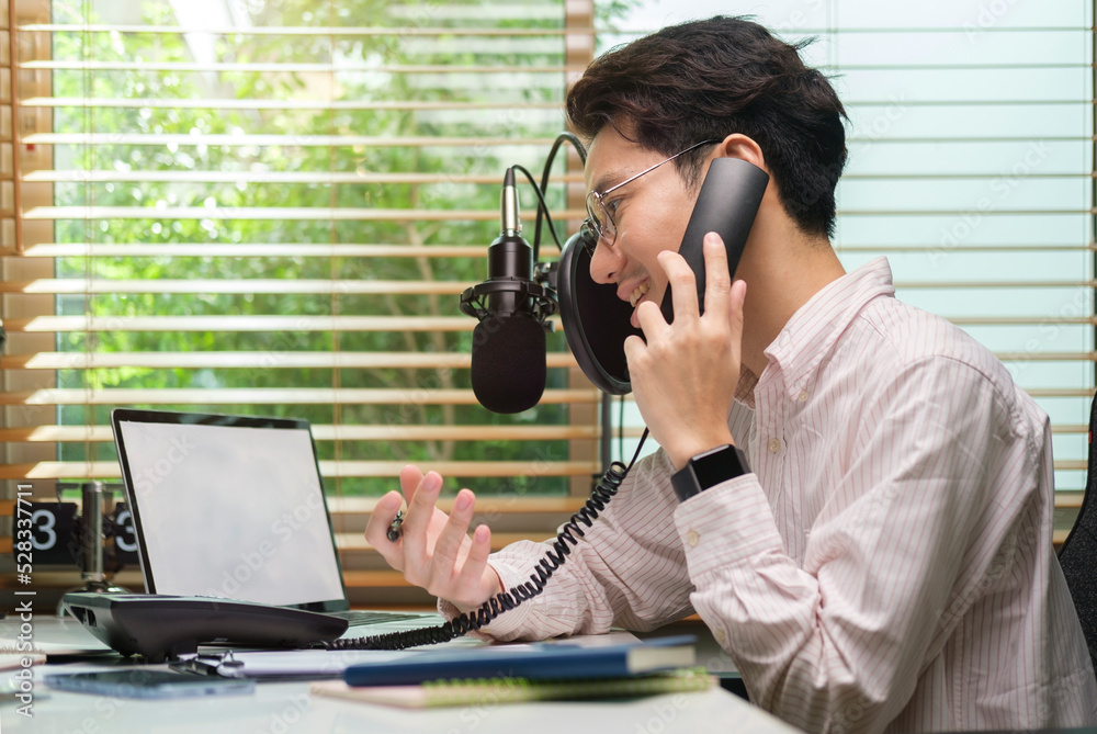 Young man vlogger recording video for social media platforms and talking on phone while sitting at home studio desk.