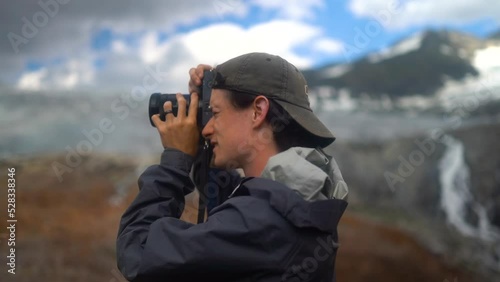 
Young caucasian male photographer lifts camera up to face to take photo of epic glacier mountain landscape