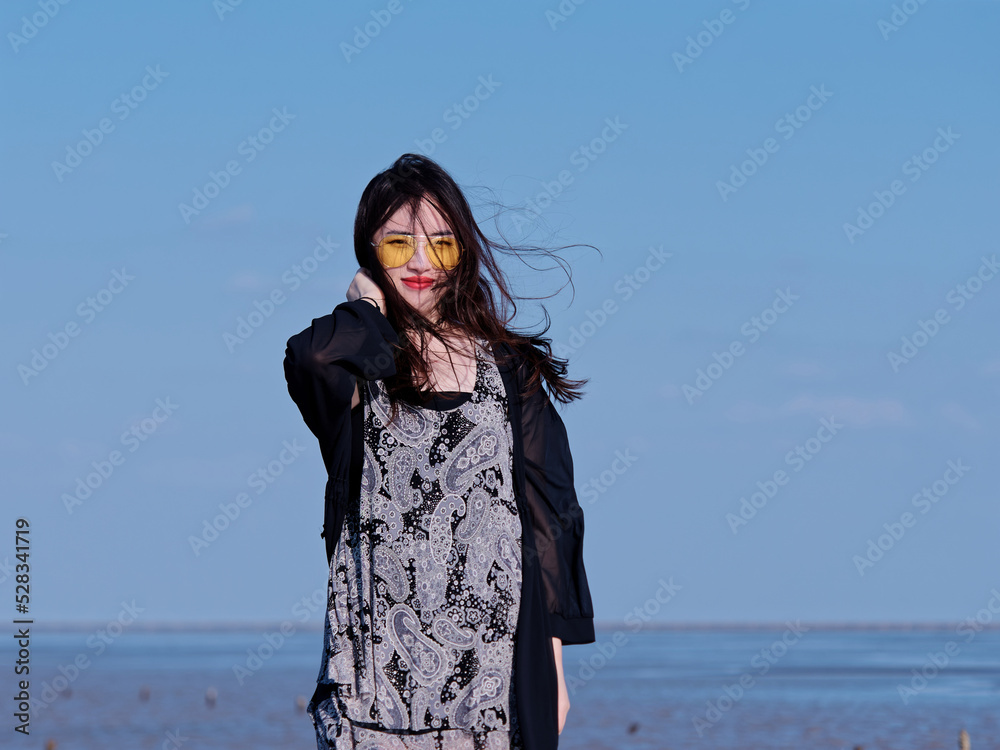 Portrait of beautiful young Chinese woman with black long hair in black dress smiling with blue sky and beach background in sunny summer day. Carefree girl enjoy her free time. Beauty lifestyle. Portrait of beautiful young Chinese woman with black long hair in black dress smiling with blue sky and beach background in sunny summer day. Carefree girl enjoy her free time. Beauty lifestyle.