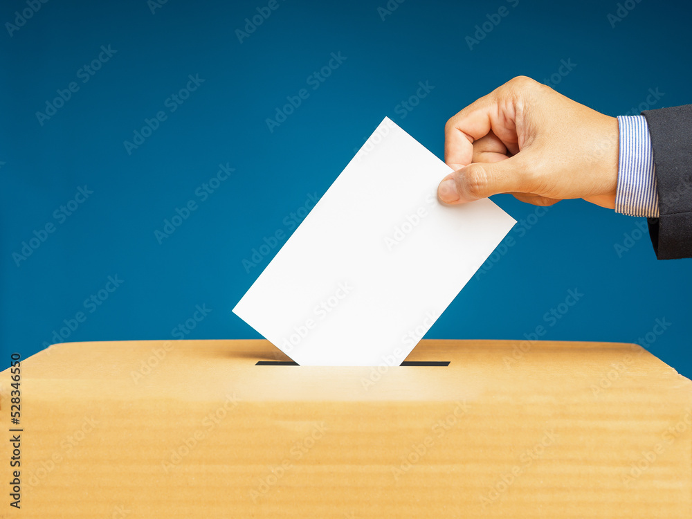 Hand voter holding ballot paper putting into the voting box at place election against a blue background Hand voter holding ballot paper putting into the voting box at place election against a blue background