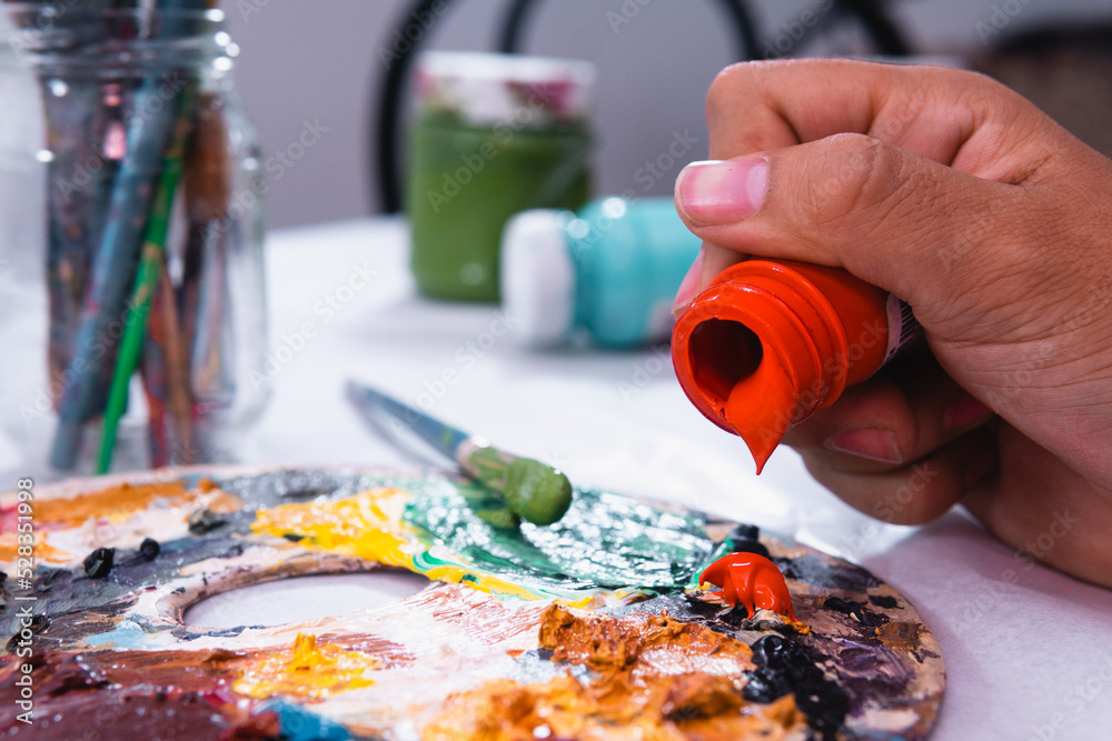 High quality photography. Paint pots of different colors, paint palette with a lot of stains and brushes in a glass jar. An artist's hand placing paint on an orange palette. High quality photography. Paint pots of different colors, paint palette with a lot of stains and brushes in a glass jar. An artist's hand placing paint on an orange palette.