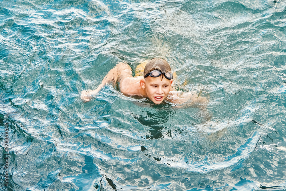 Junior schoolboy in swimming goggles enjoys holidays. Schoolkid swims in clear blue sea with smiling and excited expression on face closeup