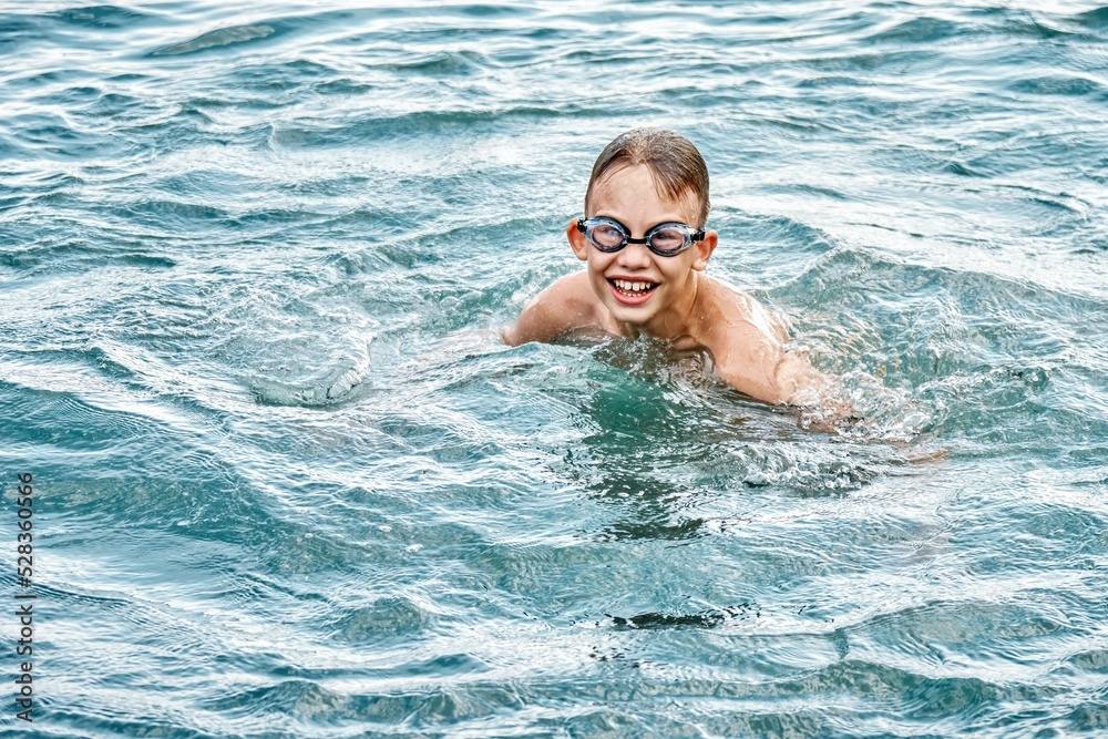 Junior schoolboy in swimming goggles enjoys holidays. Schoolkid swims in clear blue sea with smiling and excited expression on face closeup