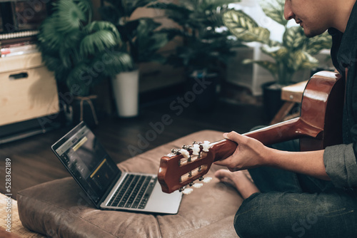 young man relax and playing guitar while sitting on sofa bed in living room at home. Music create melody song, lyrics on laptop and practice concept.
