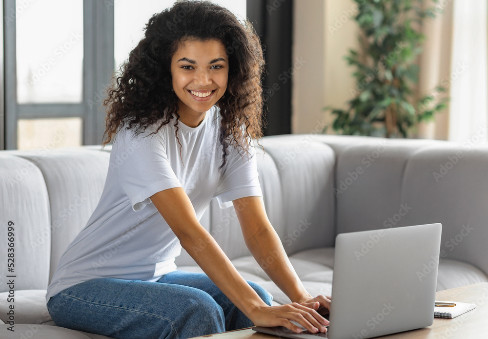 Young African American woman looking at camera and smiles. Female student or freelancer working or watching webinar using laptop sitting on at home. Online learning, distance education Young African American woman looking at camera and smiles. Female student or freelancer working or watching webinar using laptop sitting on at home. Online learning, distance education