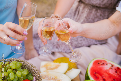 Women clink glasses with white wine or champagne at an outdoor party. Women's hands hold transparent glasses with an alcoholic drink at a picnic.Close-up. Juicy fruits in the background.