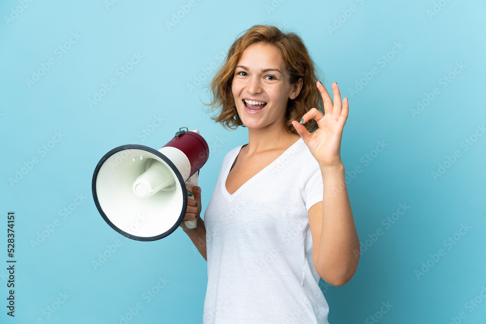 Young Georgian woman isolated on blue background holding a megaphone and showing ok sign with fingers Young Georgian woman isolated on blue background holding a megaphone and showing ok sign with fingers