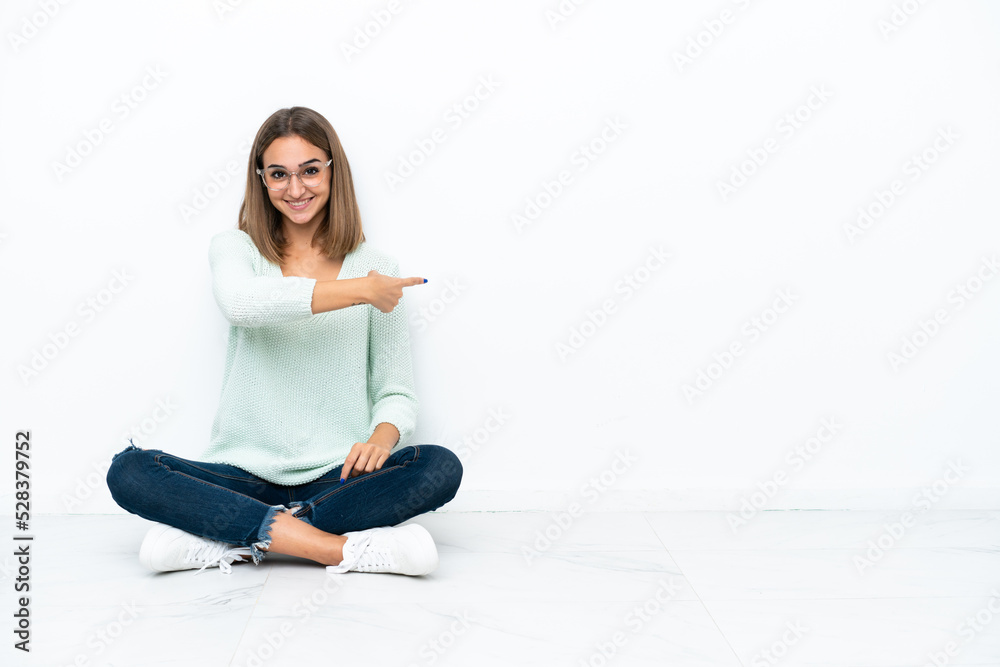 Young caucasian woman sitting on the floor isolated on white background pointing back