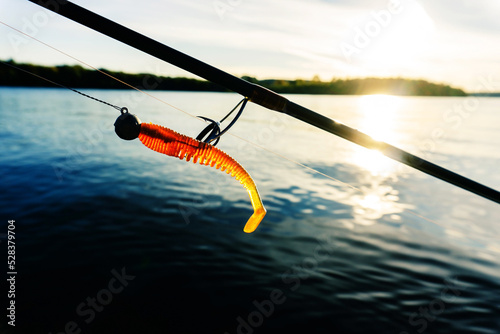 A fragment of a spinning rod and a silicone bait against the background of the river and the sun's rays. Fishing for predatory fish in the summer. Foreground