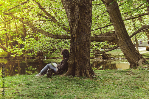 Attractive young woman reading book while sitting on grass in green public park. Springtime outdoors. Greenery unity with nature. Spend free time on open air. Education concept #528393353