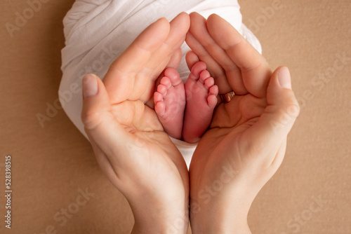 The palms of the parents. A mother hold the feet of a newborn child in a white blanket on a beige orange background.. The feet of a newborn in the hands of parents. Photo of foot, heels and toes