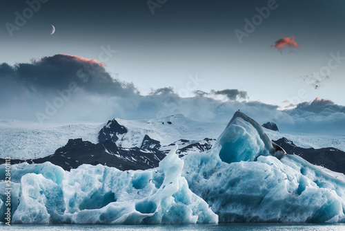Fotografie, Obraz Scenery of blue iceberg floating and the moon in Jokulsarlon glacial lagoon on d