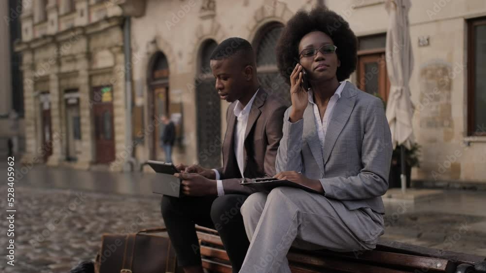 Busy afro american woman having mobile conversation while male partner sitting near on wooden bench and using digital tablet. Concept of modern gadgets and remote work. Busy afro american woman having mobile conversation while male partner sitting near on wooden bench and using digital tablet. Concept of modern gadgets and remote work.