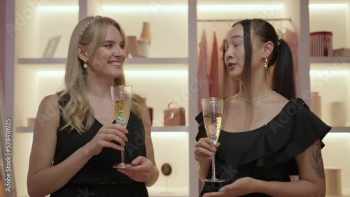 Two female friends in black dresses drinking sparkling wine standing in wardrobe and talking. Women preparing for the party. photo