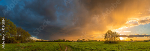 Stunning panorama of a dramatic sunset over a summertime field and forest, with heavy storm clouds in the background.