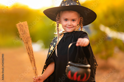 cute little blonde girl dressed in a witch costume celebrating in the park, holding a candy bowl in her hand, looking at the camera, space for text