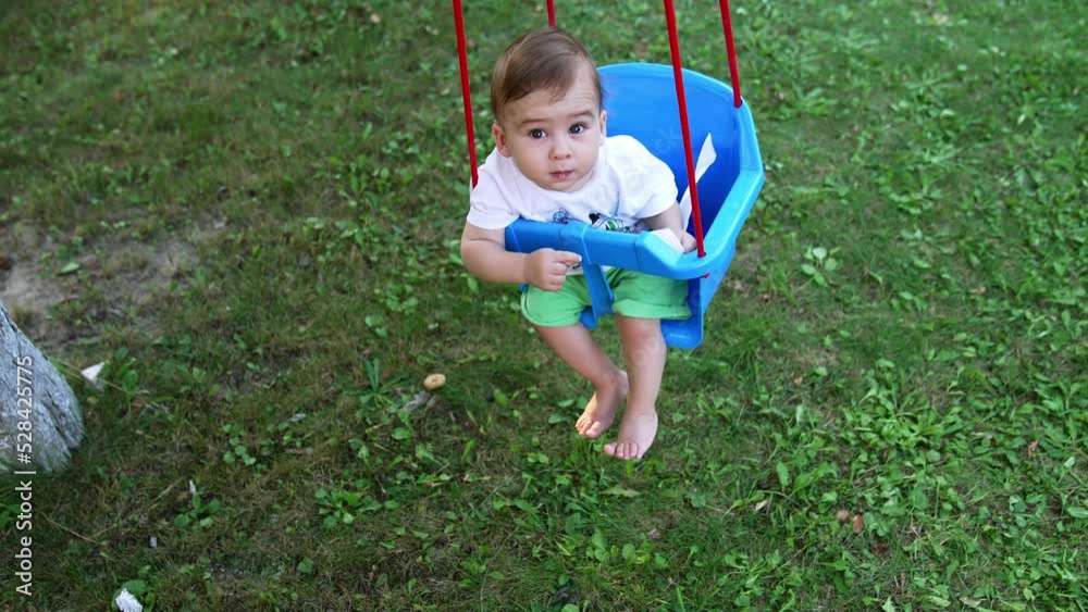 Sweet little Caucasian boy looking into camera swaying in a swing. High angle view. Green grass at backdrop. Sweet little Caucasian boy looking into camera swaying in a swing. High angle view. Green grass at backdrop.