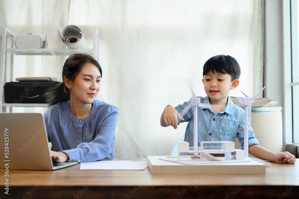 Little kid son enjoys playing with wind turbine near Young Asian mother is an energy and environmental engineer while working in home office, teaching and instilling ideas about natural energy. Little kid son enjoys playing with wind turbine near Young Asian mother is an energy and environmental engineer while working in home office, teaching and instilling ideas about natural energy.