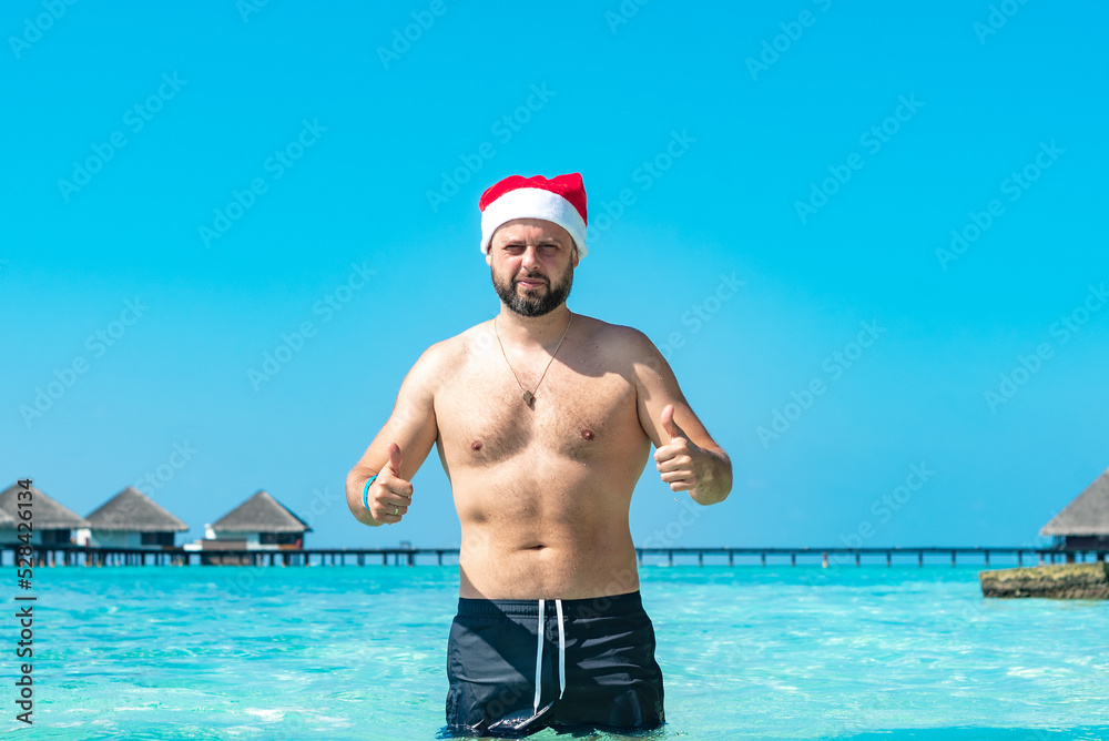 young man in Santa hat stand on near water villas in the Maldives, concept of winter holidays and luxury travel