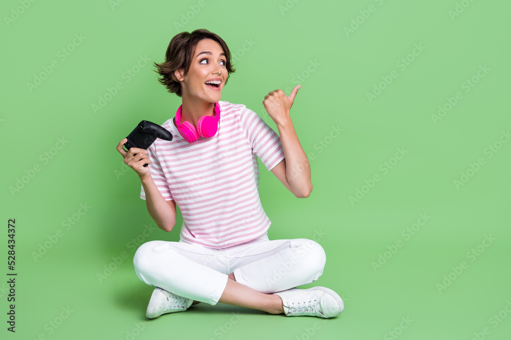 Full length photo of nice young girl point empty space playstation earphones dressed trendy striped look isolated on green color background Full length photo of nice young girl point empty space playstation earphones dressed trendy striped look isolated on green color background
