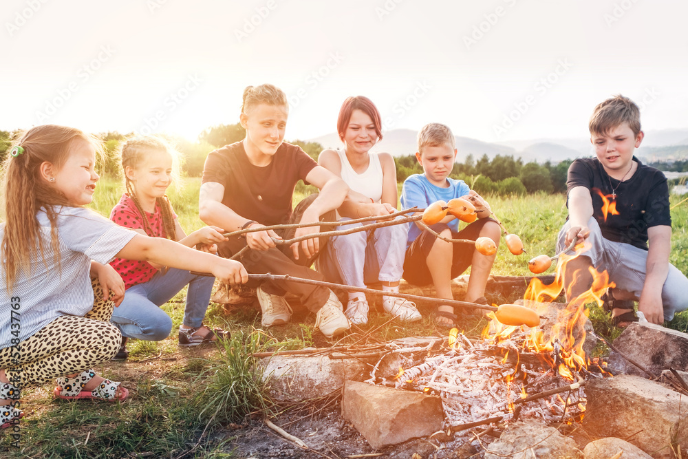 Six kids group Boys and girls cheerfully laughed and roasted sausages on sticks over a campfire flame near the green tent. Outdoor active time spending or camping in Nature concept. - obrazy, fototapety, plakaty 