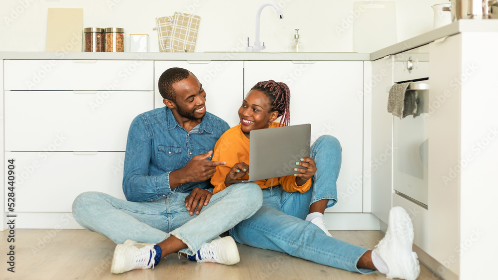 Happy black wife and husband using laptop, watch video or shopping online in kitchen interior, sitting together on floor