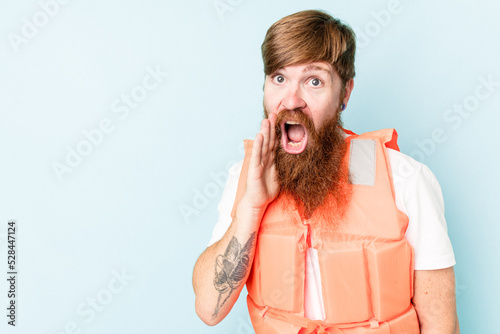 Young caucasian man wearing life jacket isolated on blue background is saying a secret hot braking news and looking aside
