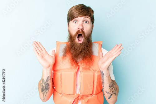 Young caucasian man wearing life jacket isolated on blue background surprised and shocked.