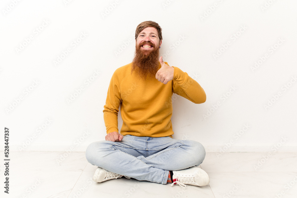 Young caucasian man sitting on the floor isolated on white background smiling and raising thumb up