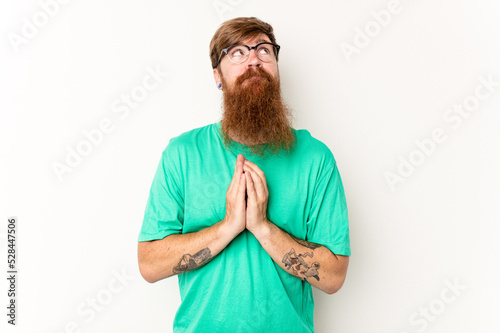 Young caucasian red-haired man isolated on white background holding hands in pray near mouth, feels confident. photo