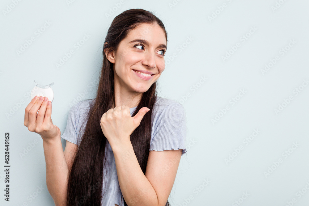 Young caucasian woman holding teeth whitener isolated on blue background points with thumb finger away, laughing and carefree. Young caucasian woman holding teeth whitener isolated on blue background points with thumb finger away, laughing and carefree.