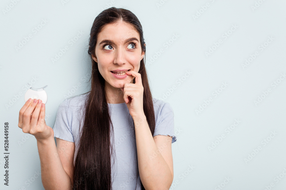 Young caucasian woman holding teeth whitener isolated on blue background relaxed thinking about something looking at a copy space. Young caucasian woman holding teeth whitener isolated on blue background relaxed thinking about something looking at a copy space.