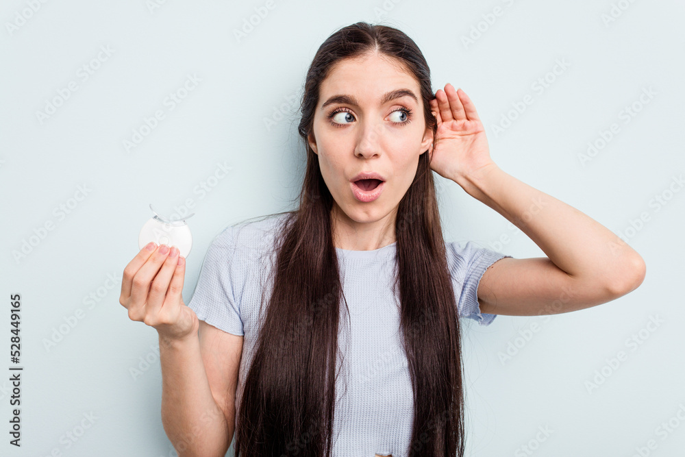 Young caucasian woman holding teeth whitener isolated on blue background trying to listening a gossip. Young caucasian woman holding teeth whitener isolated on blue background trying to listening a gossip.