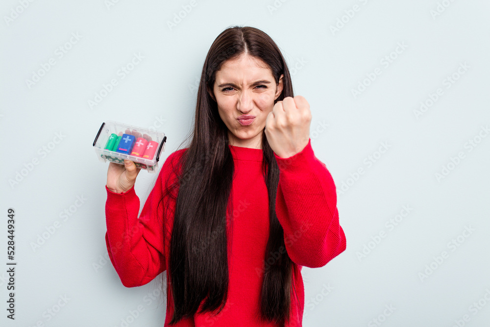 Young caucasian woman holding a batteries box isolated on blue background showing fist to camera, aggressive facial expression. Young caucasian woman holding a batteries box isolated on blue background showing fist to camera, aggressive facial expression.