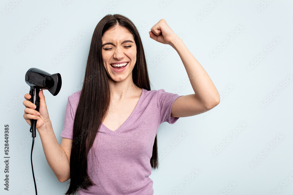 Young caucasian woman holding hairdryer isolated on blue background raising fist after a victory, winner concept. Young caucasian woman holding hairdryer isolated on blue background raising fist after a victory, winner concept.